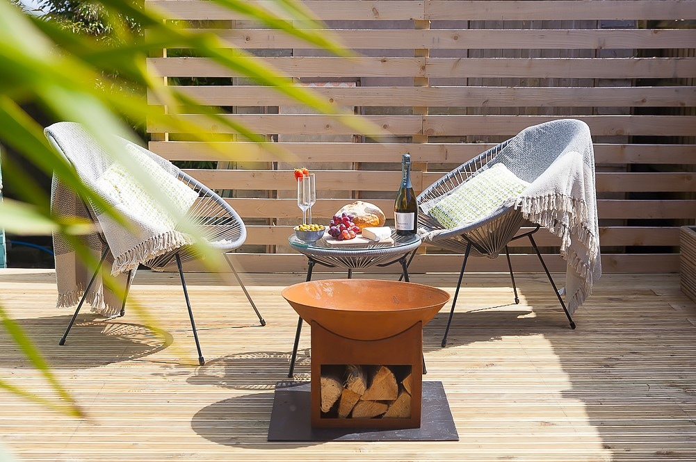 Two chair seats, draped with throws, face a small table. The table holds a bottle of champagne, glasses, and a plate with snacks. A fire pit with firewood sits nearby, surrounded by a wooden deck and privacy slats.