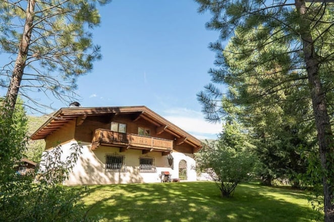 The charming exterior of the chalet is framed by tall pine trees, with a well-manicured lawn in front. A private balcony can be seen on the upper level, complementing the rustic architecture typical of European chalets.