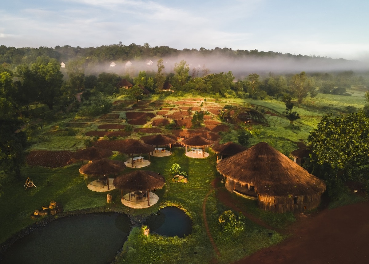An aerial view reveals a serene landscape featuring multiple thatched-roof cottages surrounded by lush greenery. A small pond reflects the open sky, while mist gently hovers over the distant hills, contributing to a calm and tranquil atmosphere.