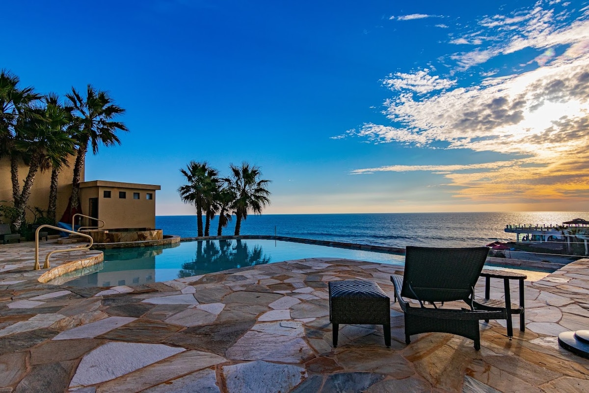 An outdoor pool area is depicted with stone flooring and lounge chairs facing the ocean. Palm trees are positioned near the poolside, while the tranquil sea and sky display varied shades of blue and soft clouds, complemented by a golden hue from the setting sun.