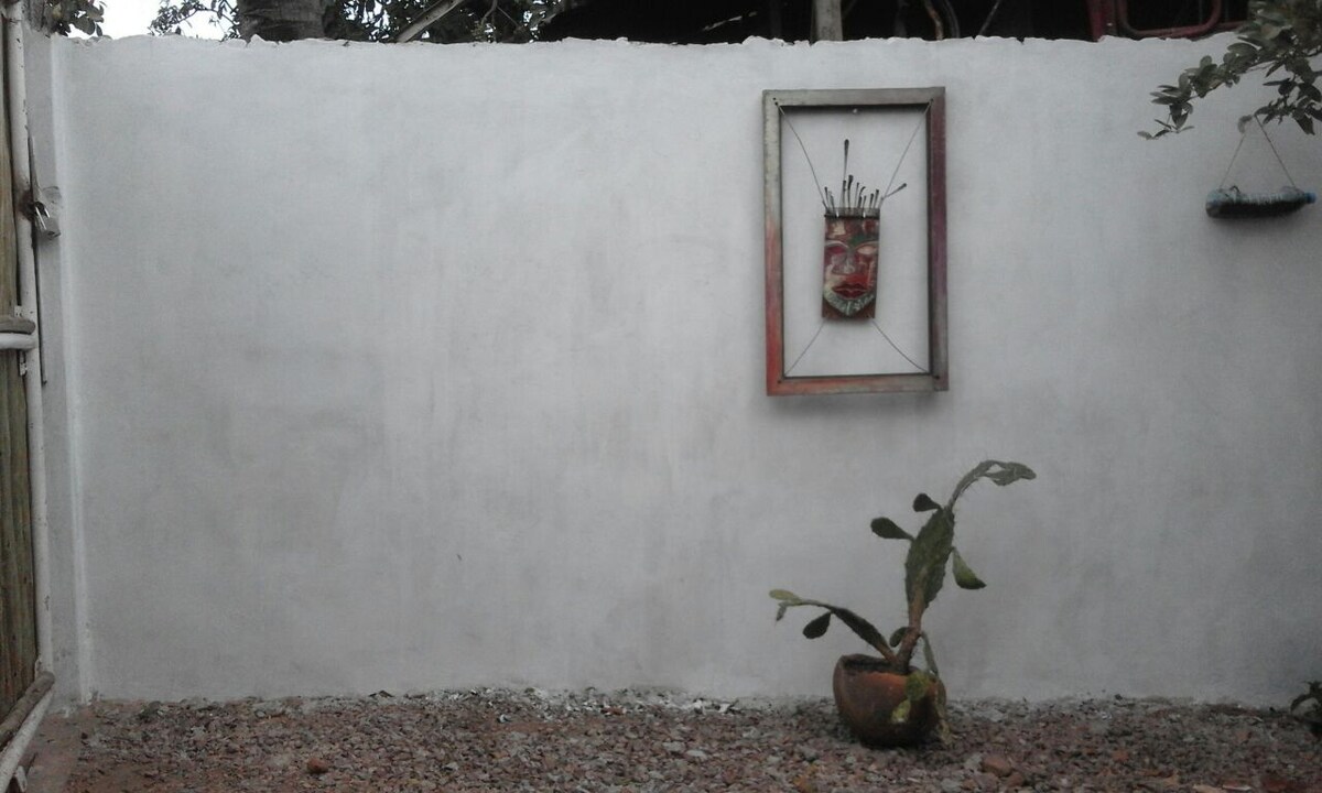 A textured white wall serves as a backdrop for a framed mask art piece, which is mounted centrally. In front, a potted plant with broad leaves sits on a rocky surface, adding a touch of greenery to the scene.