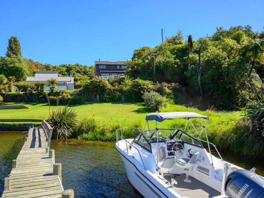 A small motorboat is docked at a wooden jetty extending into calm waters. Lush greenery surrounds the jetty and property, with a modern three-level house visible in the background, nestled among the trees. A clear blue sky creates a serene atmosphere.