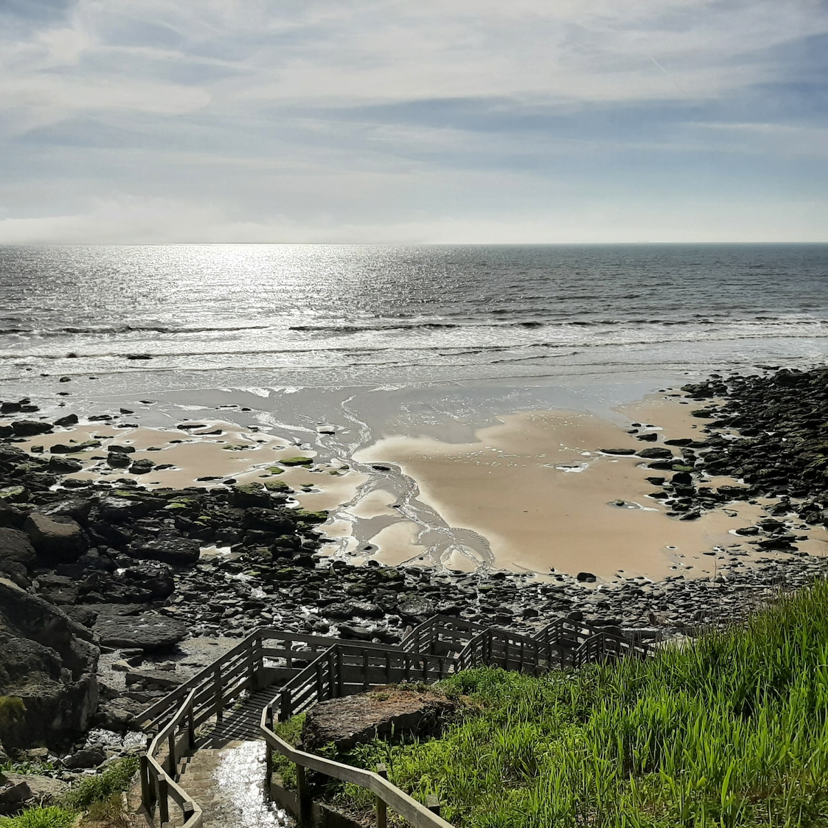 The image showcases a serene coastline with gentle waves lapping against the sandy beach. Steps made of wood lead down towards the shoreline, surrounded by green grass and rocky formations. The sunlight reflects off the water, creating a calm and tranquil scene.