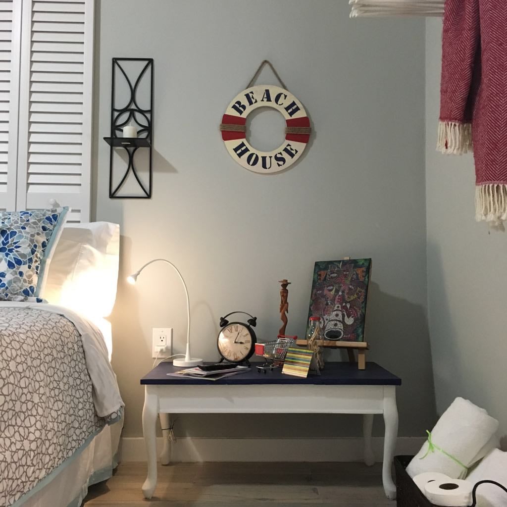 A bedside table is adorned with a small clock, a framed artwork, and an array of postcards. A decorative wall hanger displays 'BEACH HOUSE.' A modern lamp provides illumination, while a cozy blanket drapes over the edge of the bed, creating a laid-back atmosphere.