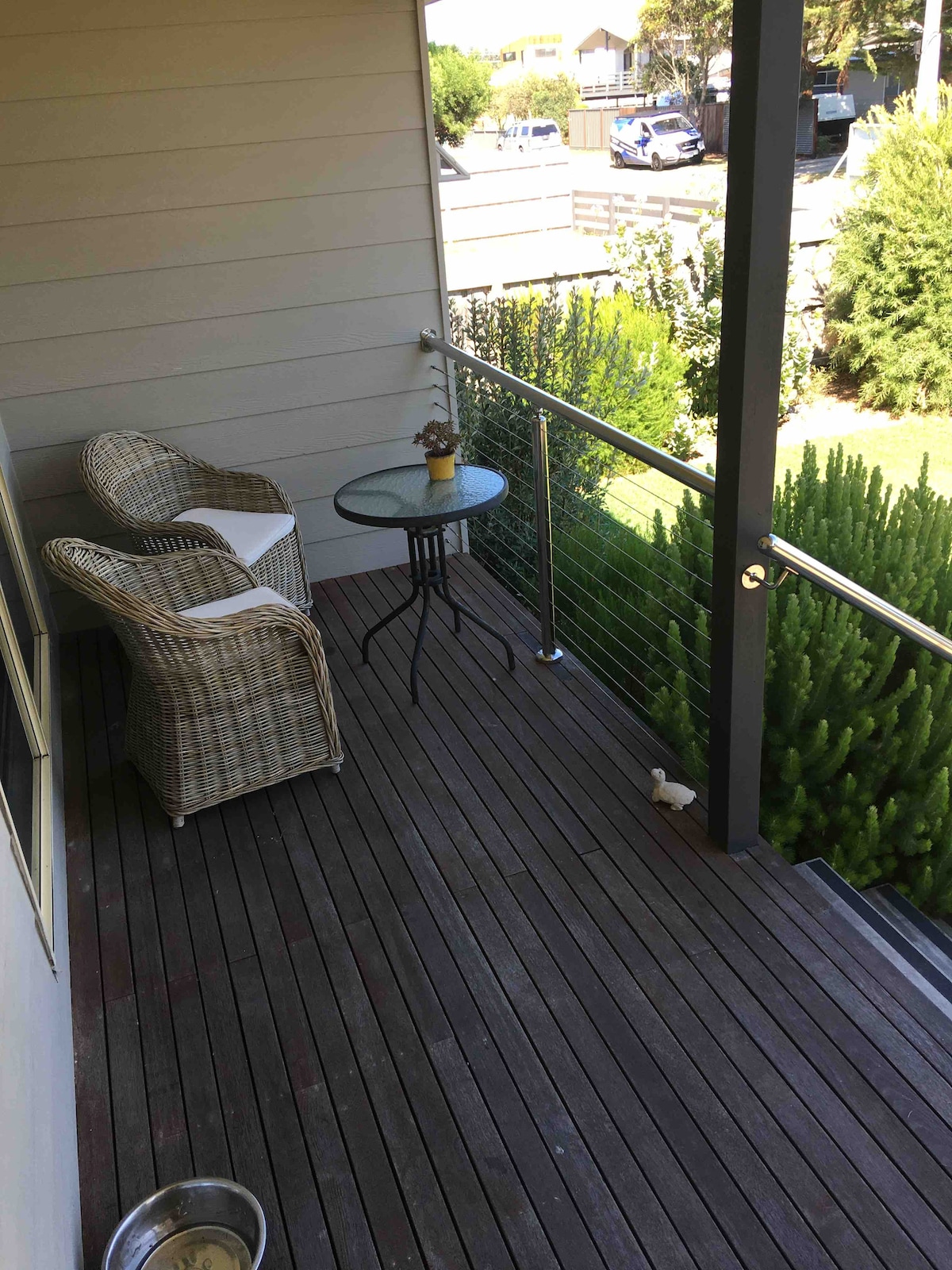 A compact outdoor balcony features a small glass table accompanied by a wicker chair. The wooden decking presents a clean and organized space, while green hedges and grass are visible just beyond the balcony, enhancing a sense of privacy.