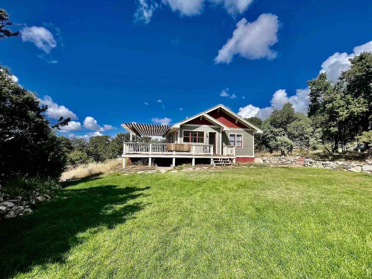 A mountain cabin is surrounded by lush green grass, with a spacious wrap-around porch showcasing a pergola. The house is set against a clear blue sky filled with scattered clouds, and tree coverage can be seen on either side.