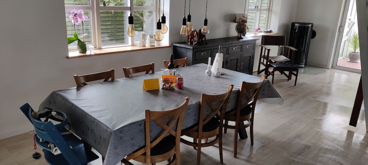 A spacious dining area is furnished with a large table surrounded by wooden chairs. A sideboard is positioned against the wall, and pendant lights hang above, providing ample illumination. Natural light enters through the windows adorned with potted plants, creating a welcoming environment.