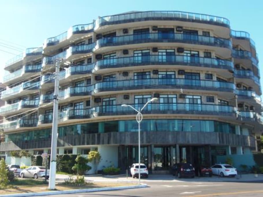Edifício visto de frente. Possui vagas para visitantes na frente do prédio.