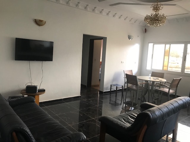 A spacious living area features black tiled flooring and white walls. A comfortable seating arrangement consists of two black leather sofas facing a wall-mounted television. A dining table with four chairs is positioned near a window, offering natural light to the space.