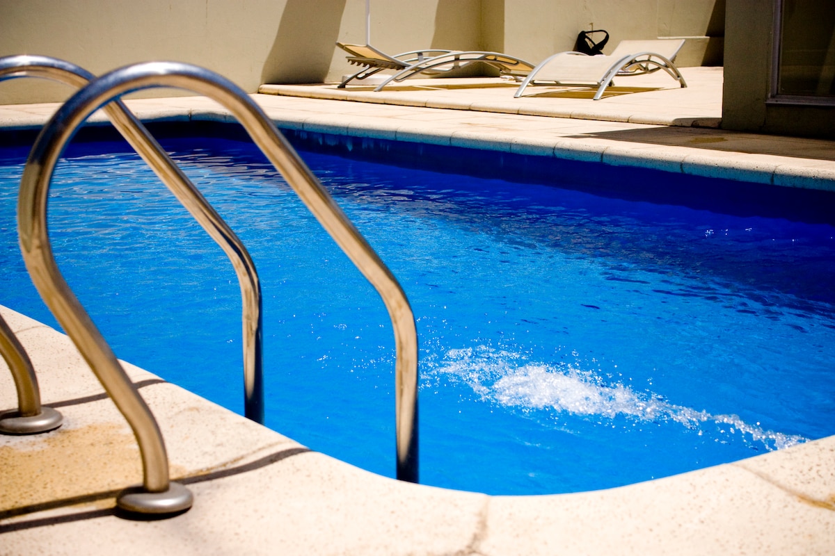 A refreshing rooftop pool is framed by a smooth stone deck. Stainless steel handrails lead into the inviting blue water. Several lounge chairs are positioned nearby, providing an ideal spot for relaxation and sunbathing.