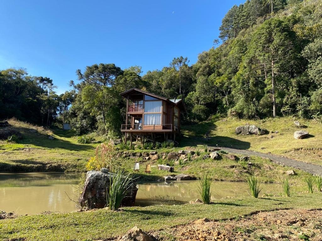 A wooden chalet is positioned on a gentle slope, surrounded by lush greenery. Large glass windows reflect natural light, providing views of the landscape. In the foreground, a tranquil pond is framed by rocks and small plantings, enhancing the serene outdoor setting.