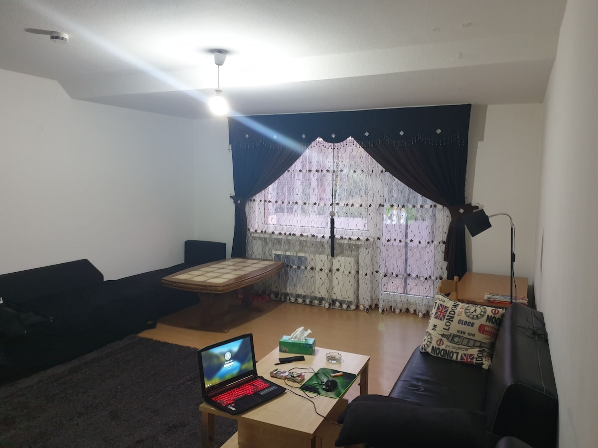 A spacious living area is showcased with a large window dressed in elegant dark curtains. A wooden coffee table sits in front of a comfortable seating arrangement, featuring two couches. A computer and decorative items are placed on a side table, enhancing the functional space.