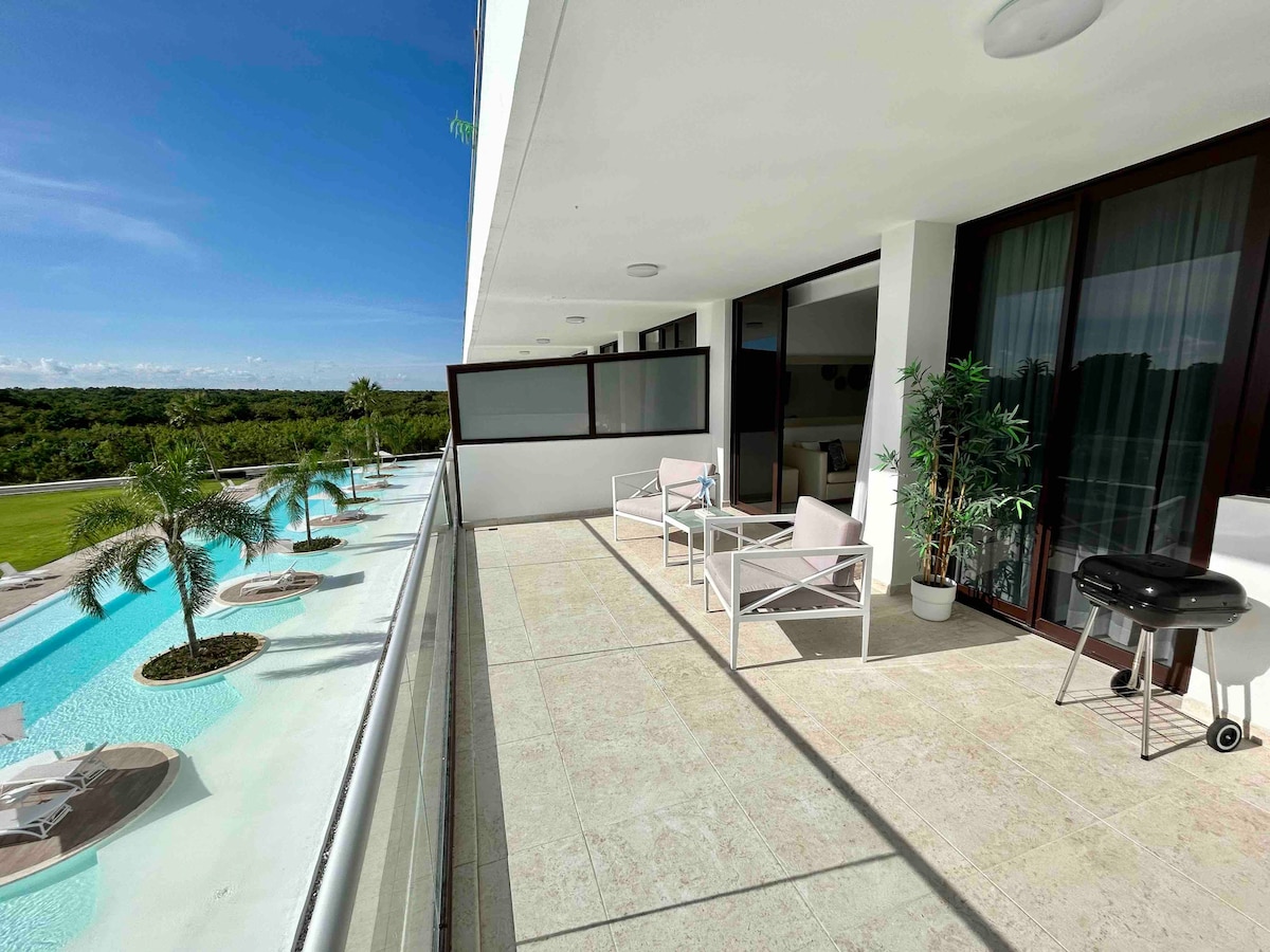 A spacious balcony overlooks lush greenery and an infinity pool. Two modern lounge chairs are positioned on the tiled floor, accompanied by a small grill. Large sliding doors provide access to the interior living space, enhancing the indoor-outdoor flow.