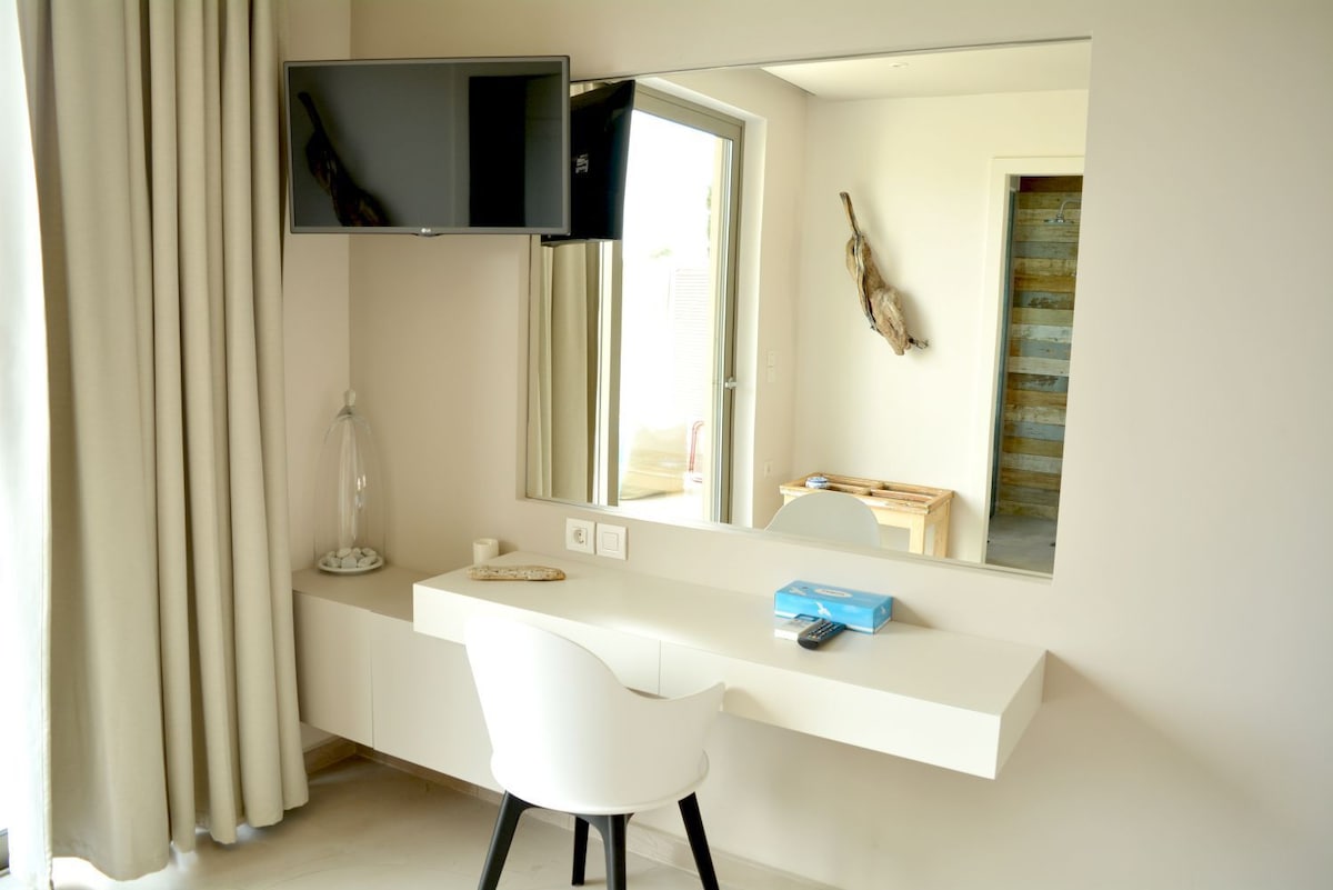 A modern workspace features a sleek white desk and a comfortable chair. A large mirror hangs above the desk, enhanced by soft lighting. A television is mounted on the wall, and natural light enters through a nearby window, complementing the minimalist decor.