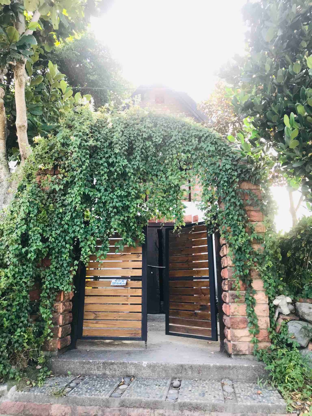 An entrance is framed by lush greenery and a wooden gate, leading into a tranquil space. The stone wall and wooden slats create a natural aesthetic. Sunlight filters through the leaves, creating a welcoming ambiance at the entryway.