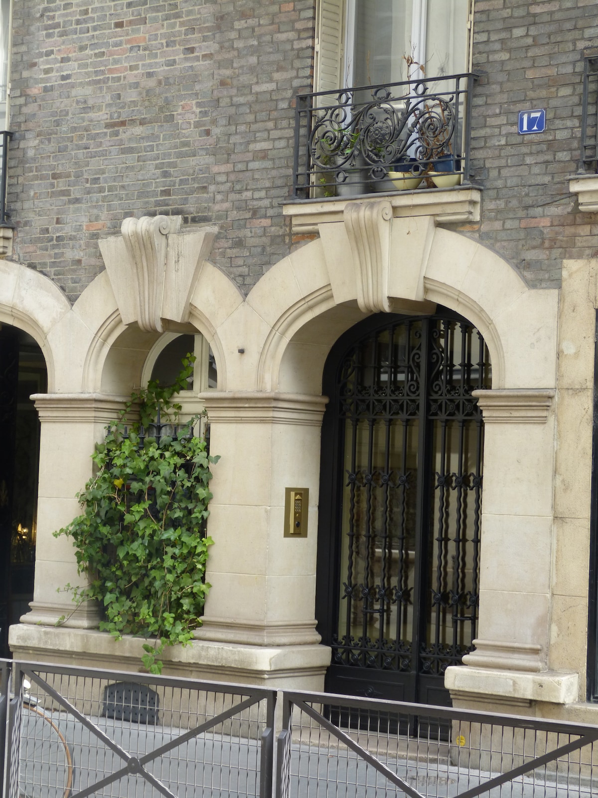 The image features a historic building entrance characterized by intricately designed stone arches and a wrought iron gate. Climbing greenery adds a natural touch to the facade. Decorative balconies are visible above, enhancing the building's architectural detail.