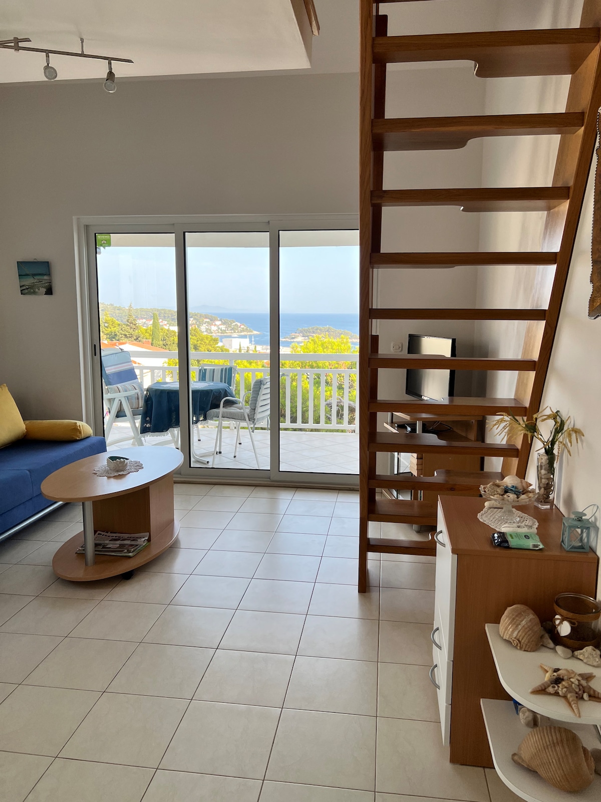 A simple living area features a blue sofa and a light wooden coffee table. A staircase leads to the upper level. Floor-to-ceiling sliding glass doors offer expansive views of the sea and access to a private balcony, filling the space with natural light.