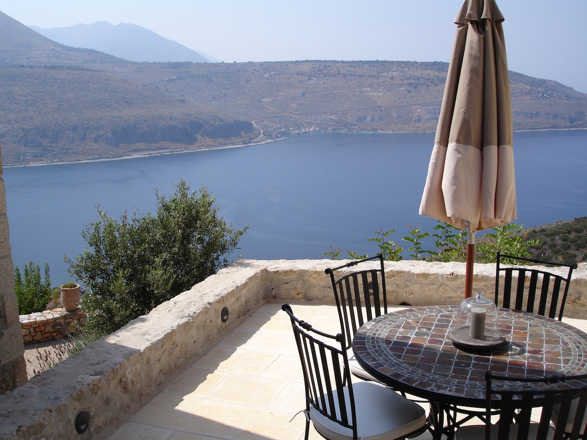 An outdoor dining area features a round table with black metal chairs, shaded by an umbrella. A coastal view of the calm blue bay is visible, framed by rolling hills in the background, offering a serene atmosphere for meals and relaxation.