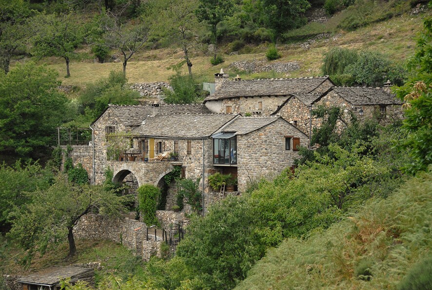 A rustic stone building is nestled among lush greenery, showcasing traditional architecture with a sloped roof and wooden details. Surrounding trees and shrubs provide a serene natural landscape, creating a peaceful retreat. The structure features both covered and open areas, inviting a connection with the outdoors.