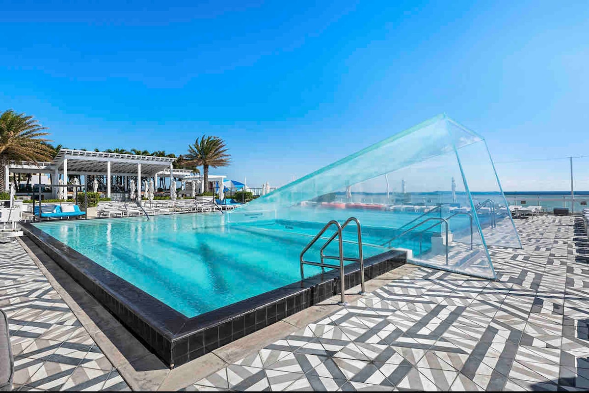 A spacious pool area is framed by a modern tile design, featuring a unique transparent edge that blends with the sky. Palm trees provide shade nearby, while lounge chairs are positioned around the pool, inviting relaxation with ocean views in the background.