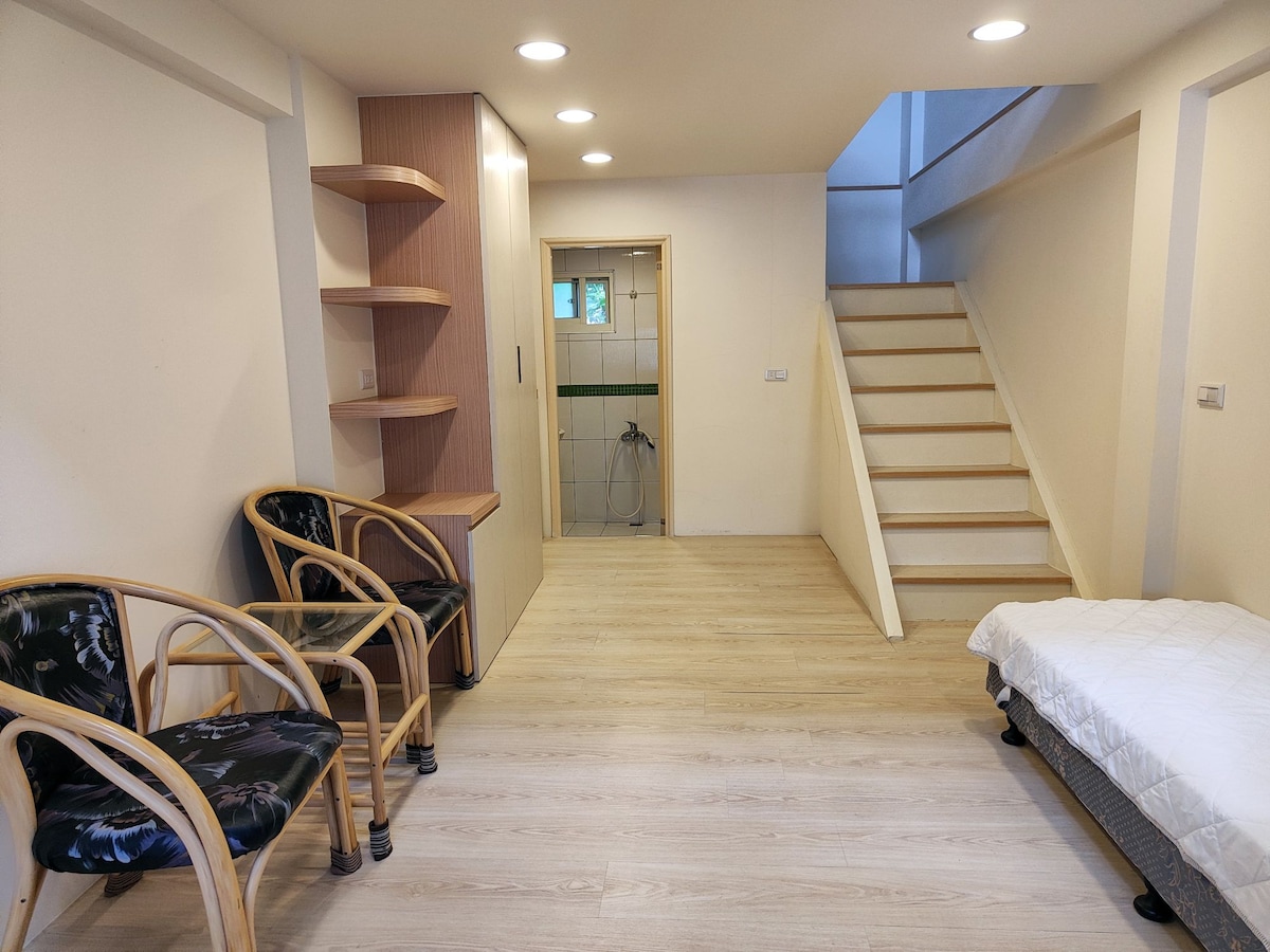 A neutrally designed interior space features light wooden flooring and minimalist furnishings. Two woven chairs are positioned near a wooden shelf, while a twin bed is located to the right. Soft lighting from recessed ceiling lights enhances the welcoming atmosphere. A staircase leads to the upper level.