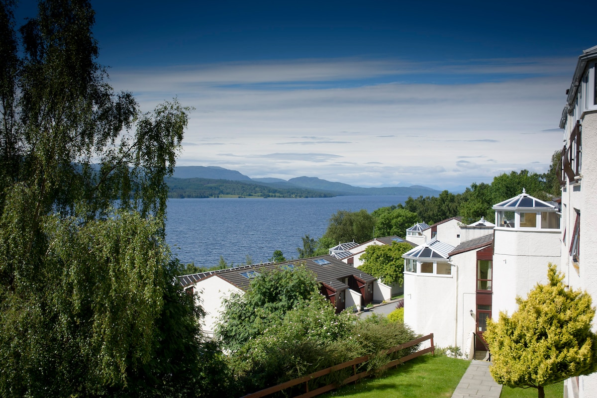 A scenic view overlooking Loch Rannoch is presented, with lush greenery and well-kept gardens in the foreground. The lodges are positioned along the water's edge, against a backdrop of distant hills under a vast sky, offering a serene and inviting atmosphere.