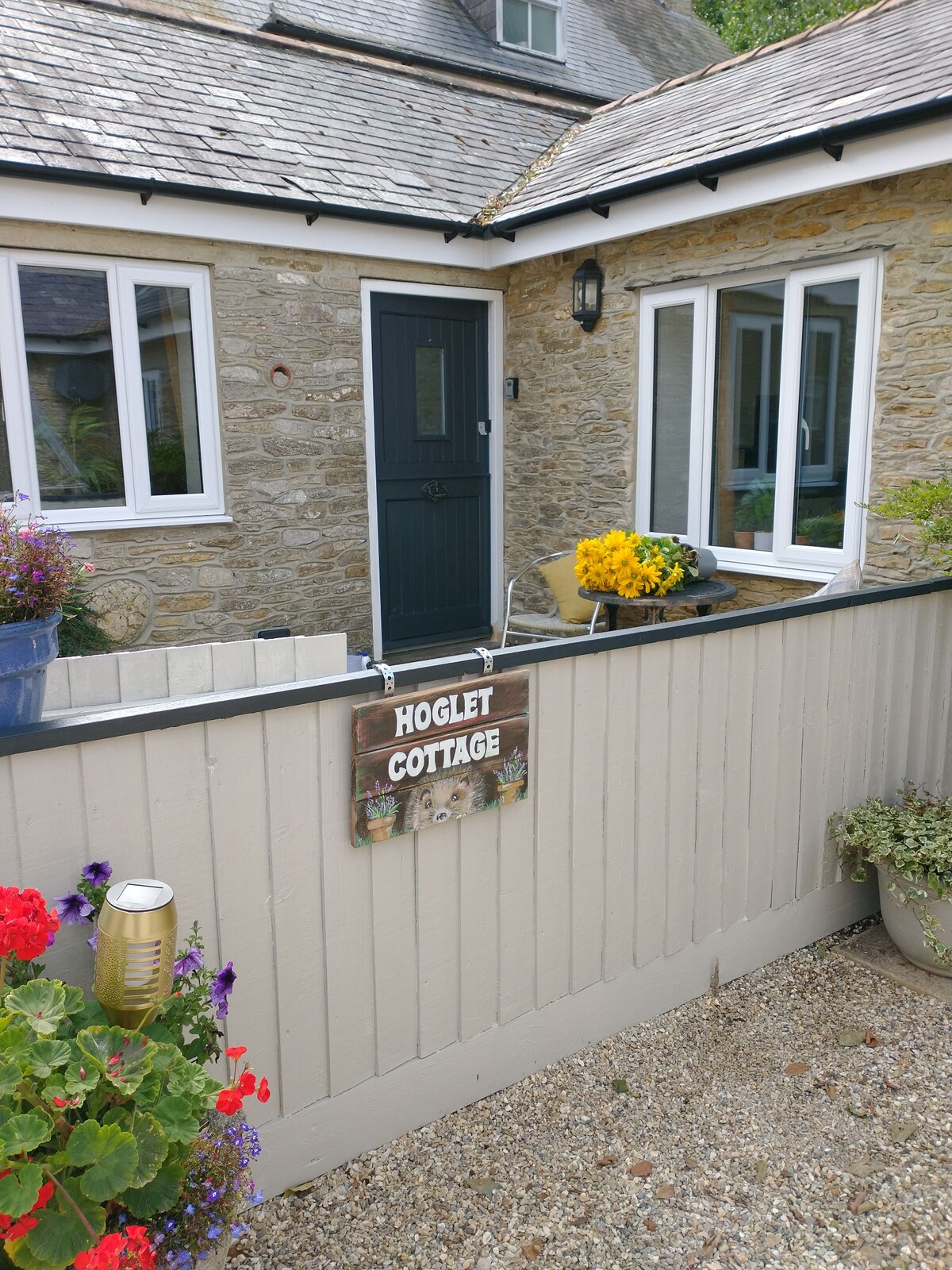The entrance of Hoglet Cottage is framed by a low wooden fence, with a stone exterior visible. A wooden sign with the name 'Hoglet Cottage' hangs near the door. Colorful flowers add vibrancy to the scene, creating an inviting entryway.