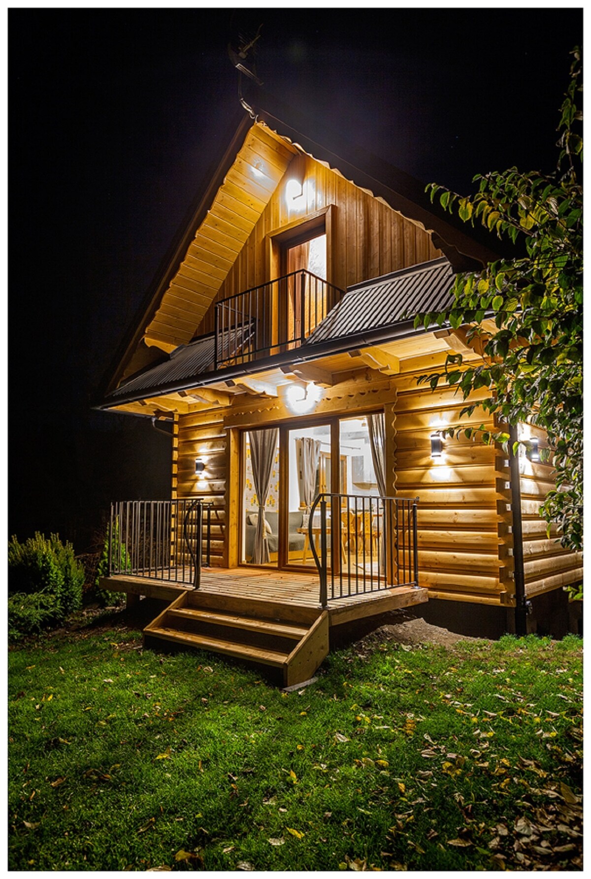 A charming wooden house features warm lighting illuminating the exterior. A small porch with stairs leads to the entrance, framed by manicured green grass and surrounding foliage. The upper balcony is adorned with elegant railing, enhancing the inviting appearance of the structure at night.