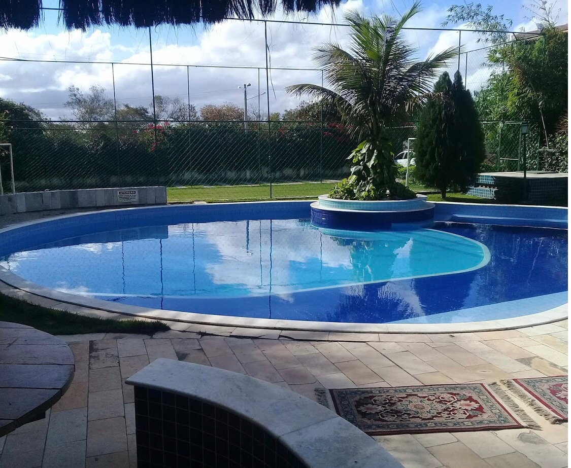 A tranquil swimming pool is surrounded by lush greenery, with palm trees and manicured shrubs enhancing the natural setting. The water's surface reflects the sky and nearby tennis courts. Paved stone flooring and a decorative rug add to the inviting surroundings.