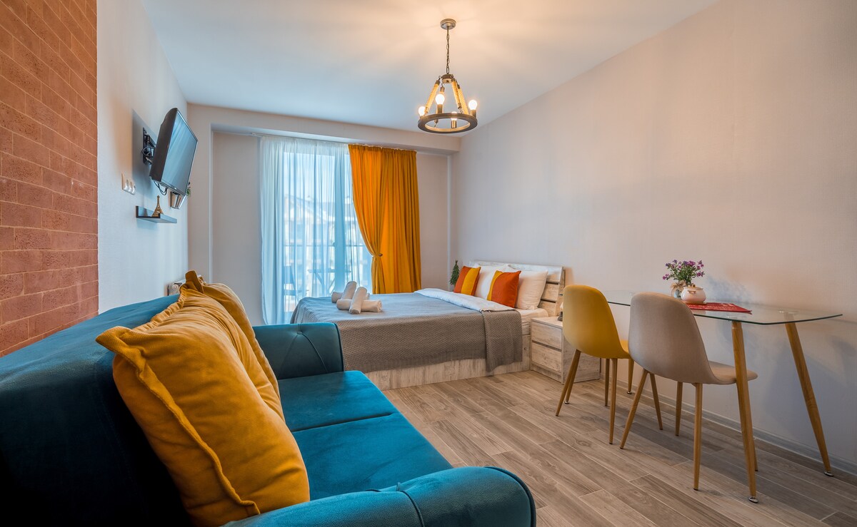 A studio apartment includes a queen bed with decorative pillows and a cozy sofa in teal. A dining table with modern chairs is set for two. Natural light enters through a large window with bright orange curtains, enhancing the inviting atmosphere. A flat-screen TV is mounted on the wall.