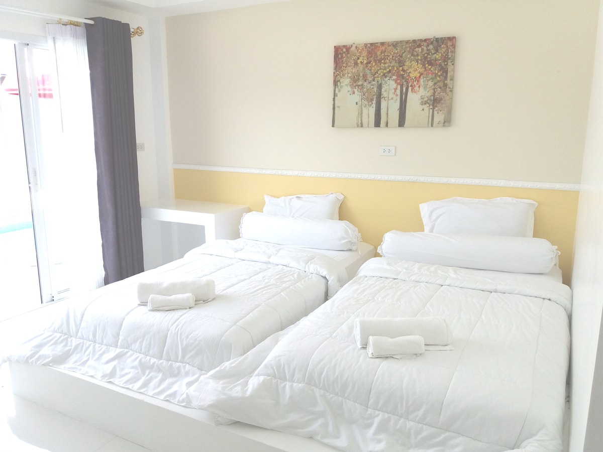 A bright sleeping area features two spacious beds with white linens and fluffy white pillows. A piece of wall art depicting trees adds a touch of color. A large window allows natural light to flow in, with sheer curtains softly framing the view.