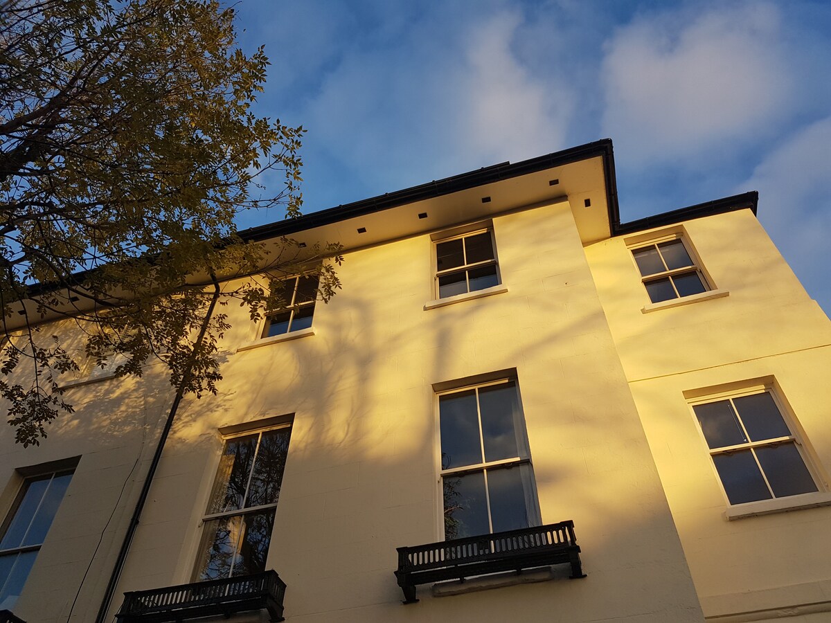 The exterior of a period building is visible, featuring a cream-colored facade with large windows. Soft shadows from nearby trees are cast upon the wall, accentuating the architectural details. The sky above is partially cloudy, creating a calm backdrop.