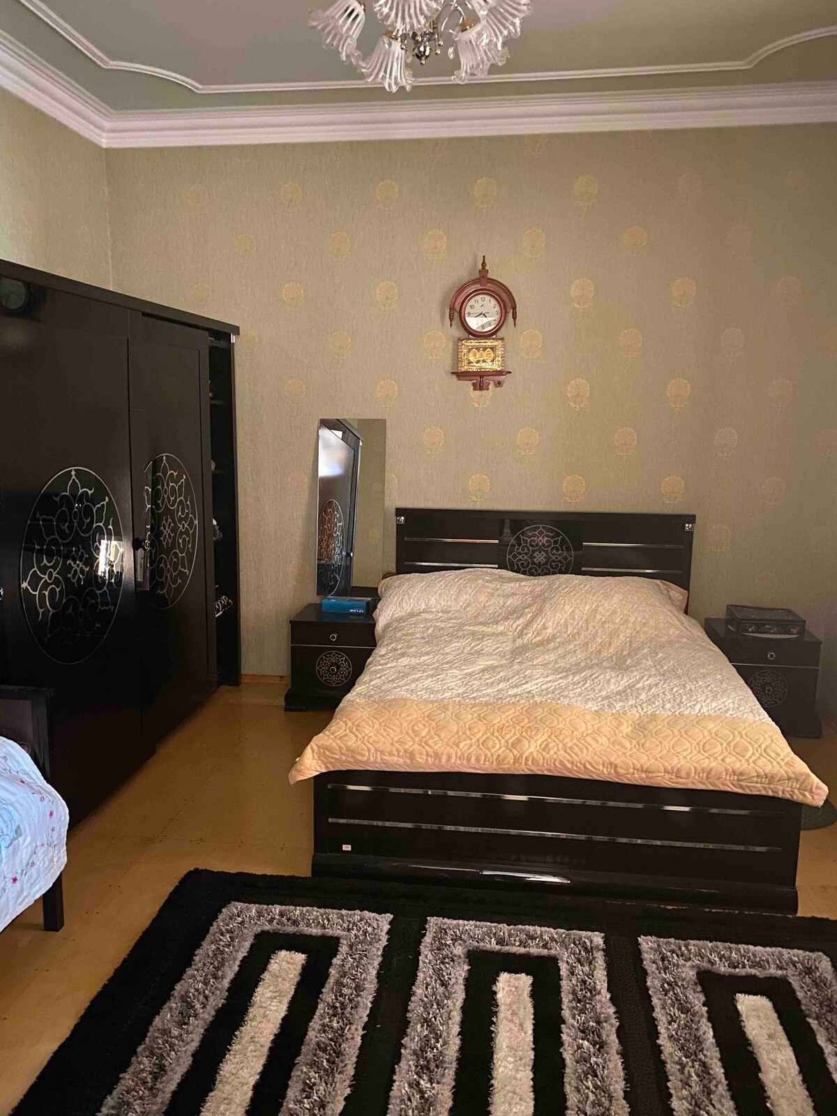 A cozy room features a dark wooden bed adorned with a light-colored quilt. A large mirror and matching nightstands complement the setting. Decorative wall art and a traditional clock enhance the space, while a plush rug adds comfort to the wooden floor.