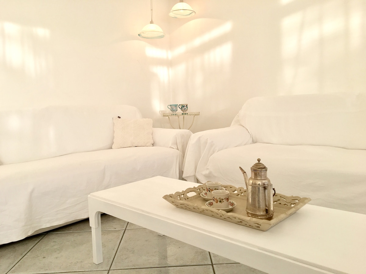 A bright living area is adorned with two white sofas positioned around a white coffee table. A decorative tray with a teapot and cups sits on the table, while natural light streams in, casting soft shadows across the walls.