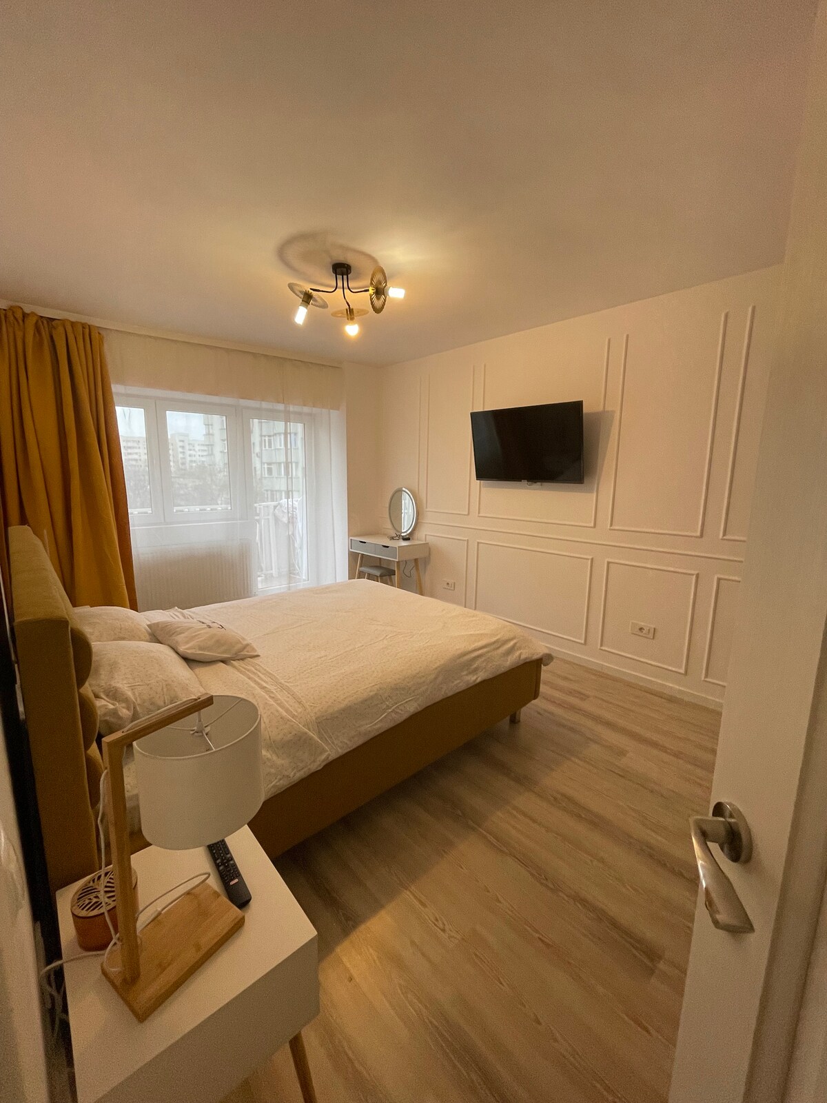 A bright bedroom is furnished with a spacious bed and a stylish wall-mounted television. Large windows allow natural light to fill the space, complemented by soft yellow curtains. A mirrored vanity and minimalist bedside lamp enhance the room's modern aesthetic.