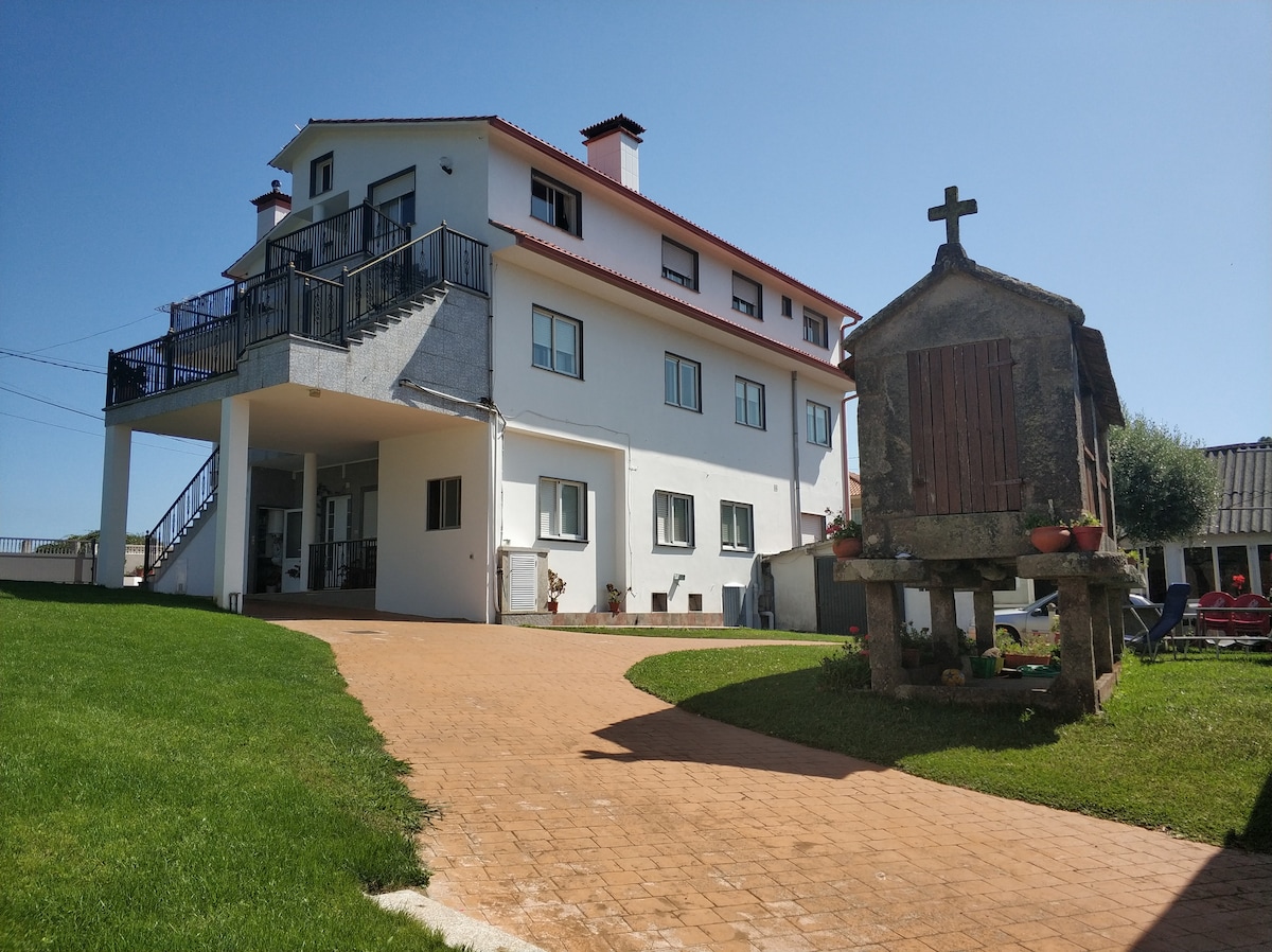 The exterior of the house features a two-story structure with a white facade and a red roof. A well-maintained garden with natural grass is visible, alongside a traditional stone granary and outdoor seating options. Clear blue skies enhance the welcoming atmosphere.