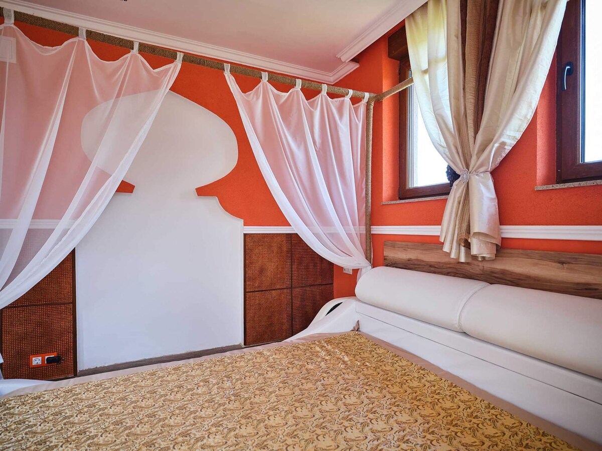A cozy sleeping area features a spacious bed with a decorative golden bedspread. Sheer white curtains frame the windows, allowing natural light to enhance the warm orange walls. Wooden accents provide a natural touch to the overall design.