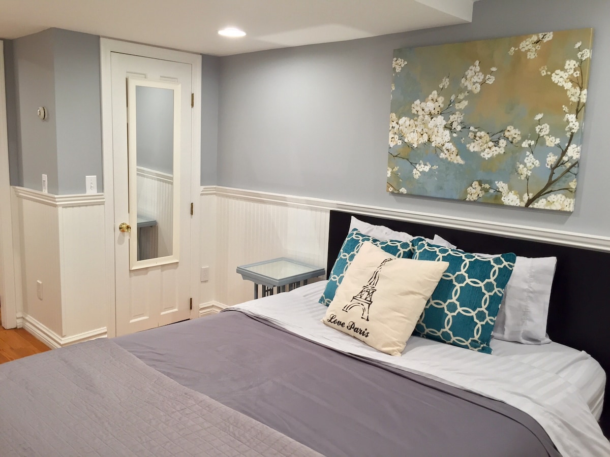 A cozy bedroom is featured, showcasing a spacious bed dressed in neutral linens. A decorative pillow with an Eiffel Tower design adds a personal touch. A floral wall art piece stands above the bed, complemented by a bedside table with a glass surface, and a mirror door reflects light.