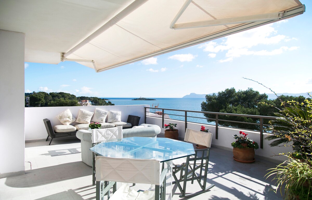 A spacious terrace features a dining table paired with chairs and a lounge area furnished with comfortable seating. Potted plants add greenery, while the expansive view of the sea and distant coastline is framed by clear blue skies and soft clouds.