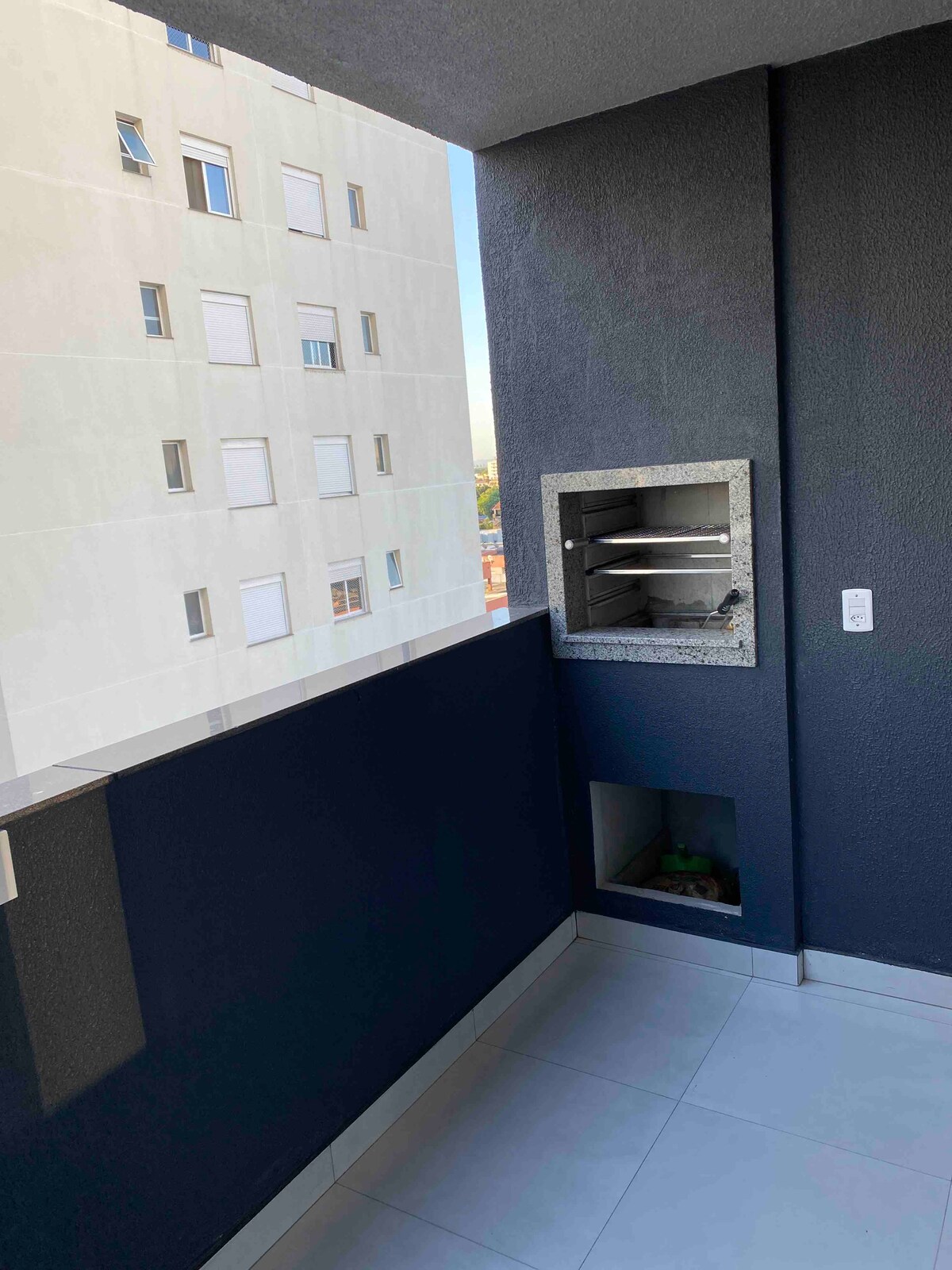 A balcony space is shown with a built-in barbecue grill positioned against a dark wall. The view reveals adjacent residential buildings, with light filtering through the nearby windows. The ground is tiled, and a seamless railing enhances the openness of the area.