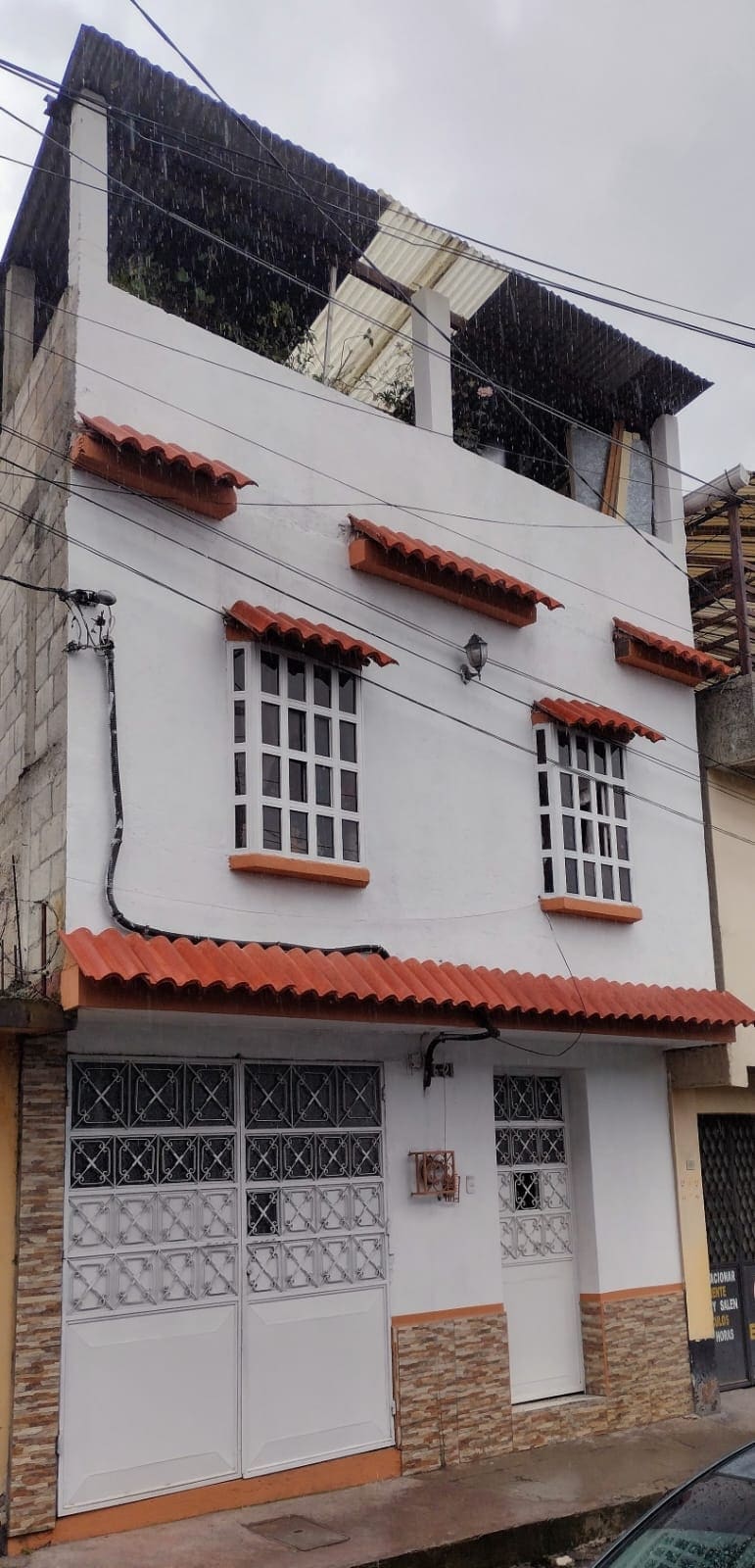 A modest two-story white house is depicted, featuring a tiled roof and decorative window frames. Vertical bars accentuate six windows on the facade, and a white door is positioned near the entrance. Above, an open terrace area is visible.