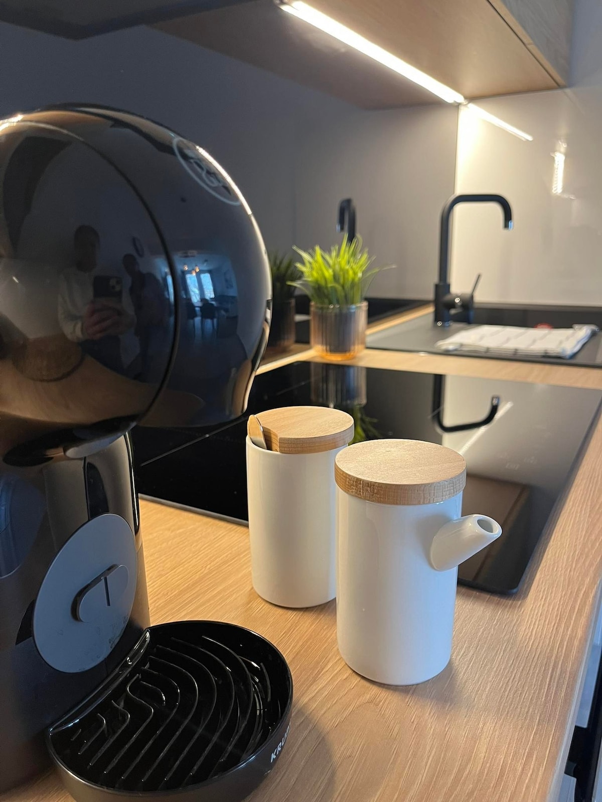 A modern kitchen setup features a sleek coffee machine alongside two white containers with wooden lids. A small plant adds a touch of greenery, while a black stovetop is visible in the background, showcasing a stylish and functional design.