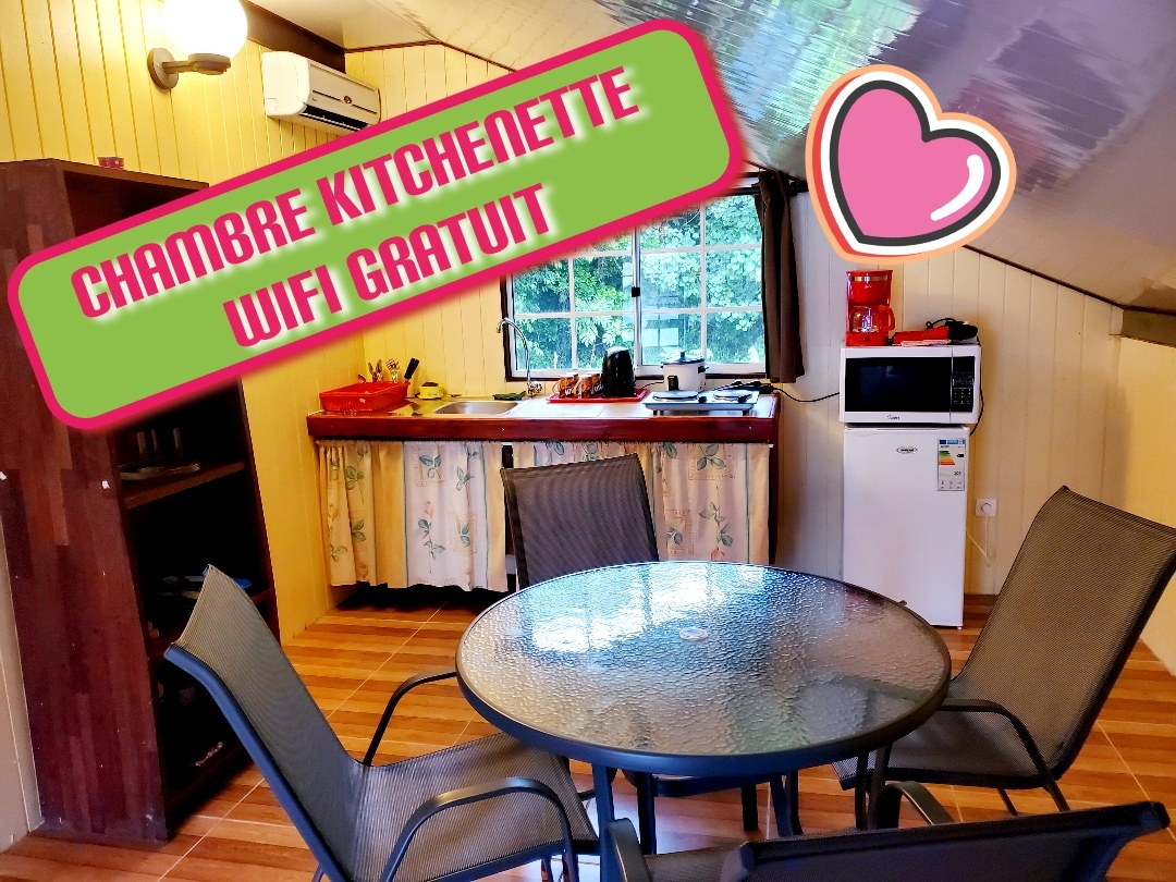A well-lit kitchenette area features a small table with chairs and various cooking appliances. A microwave and coffee maker are visible. Cabinets with patterned curtains provide storage. Natural light streams through the window, enhancing the space's inviting ambiance.