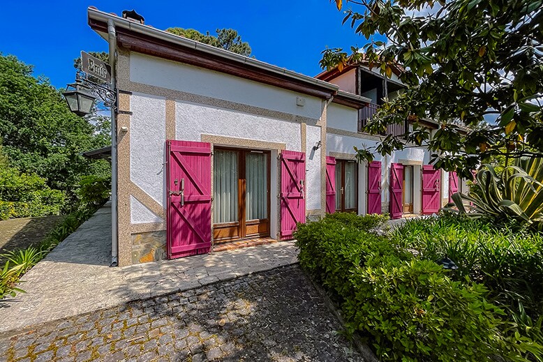 The exterior of the Casa EMIR showcases a charming facade with bright pink shutters and large windows. Surrounding greenery enhances the welcoming entrance, while stone paving leads to the door. The building is complemented by adjacent architecture, contributing to the tranquil setting.