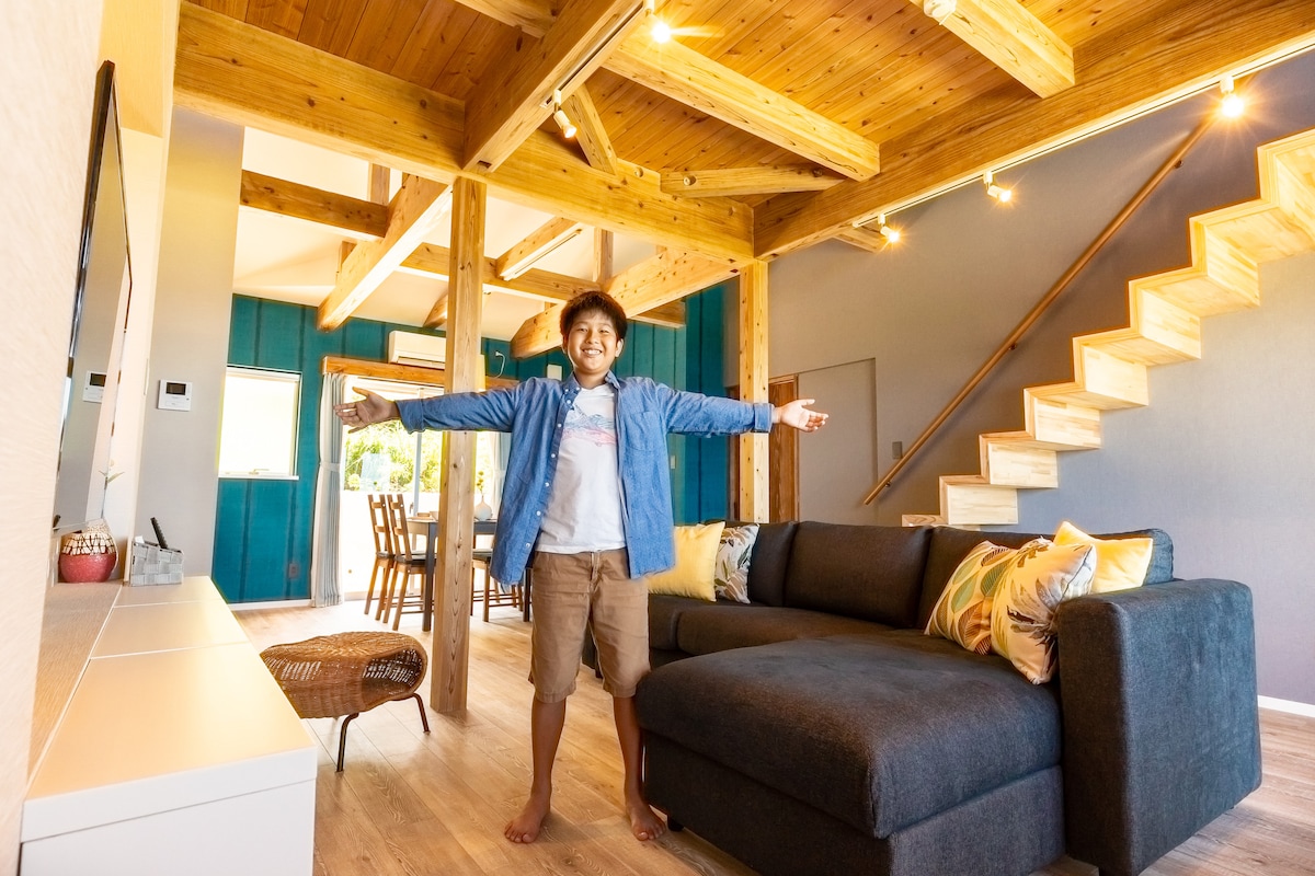 A spacious living area features a large dark sofa with decorative cushions, framed by exposed wooden beams and a high ceiling. A staircase with wooden steps leads to an upper level, while a dining area is visible in the background.