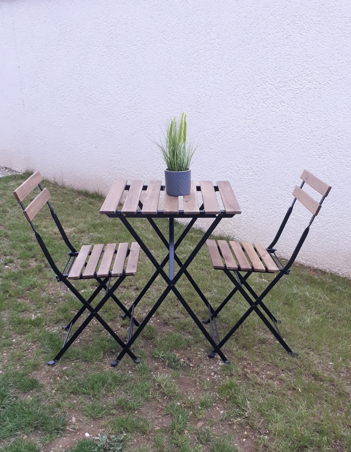 A small, foldable outdoor table is positioned on a grassy area, accompanied by two matching folding chairs. A potted plant sits in the center of the table, adding a touch of greenery. The backdrop consists of a plain, light-colored wall.