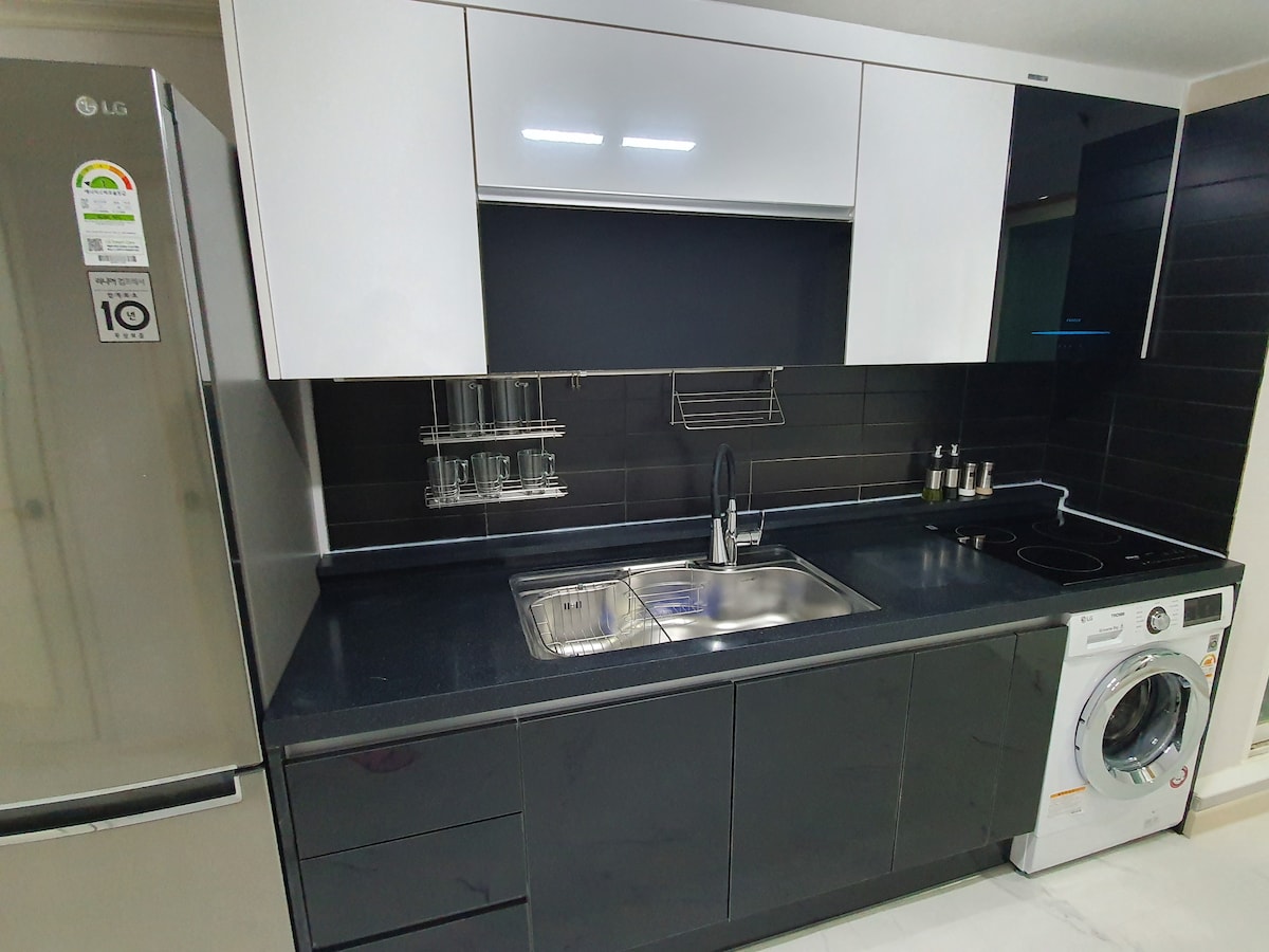A modern kitchen is presented, featuring sleek black cabinetry and a stainless steel refrigerator. A stainless steel sink is positioned next to a four-burner stovetop and a washing machine. Wall-mounted storage is visible above the countertop, providing easy access to kitchen essentials.