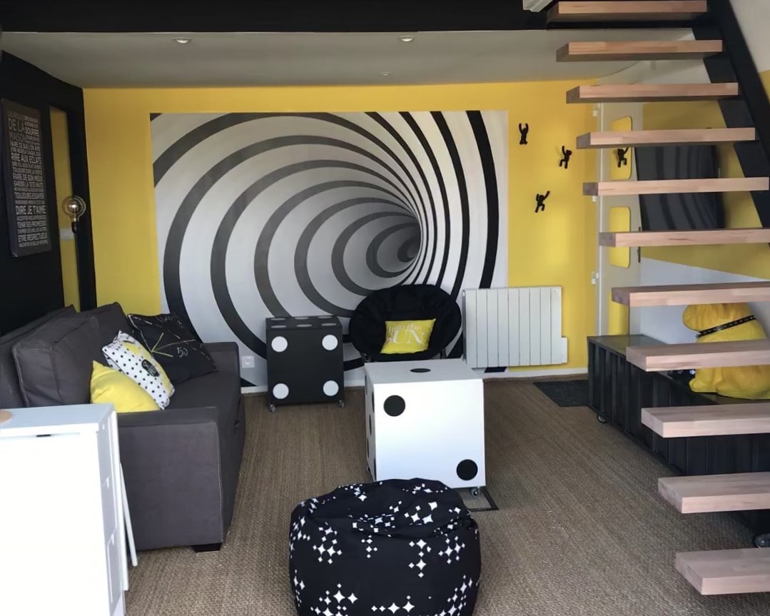 A modern living area is highlighted by a striking black and yellow spiral wall art. A grey sofa sits beside playful black and white accent tables. A cozy bean bag chair and two poufs provide additional seating, while a staircase leads to the upper level.