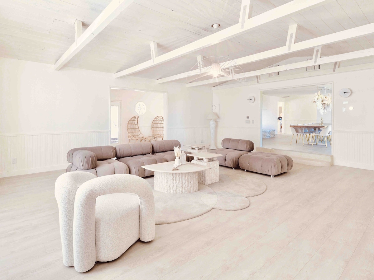 A spacious living area features soft, curved furniture in neutral tones, including a large sectional sofa and unique accent chairs. A round, textured coffee table sits on an organic-shaped rug, enhancing the open layout illuminated by natural light from surrounding areas.