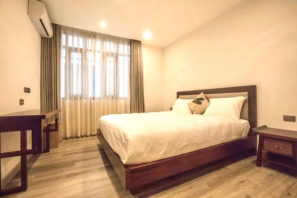 A bright and airy bedroom features a spacious bed with a decorative pillow. Natural light filters through large windows draped with light curtains. A wooden desk and a bedside table complement the minimalist decor, while neutral tones create a calm environment.